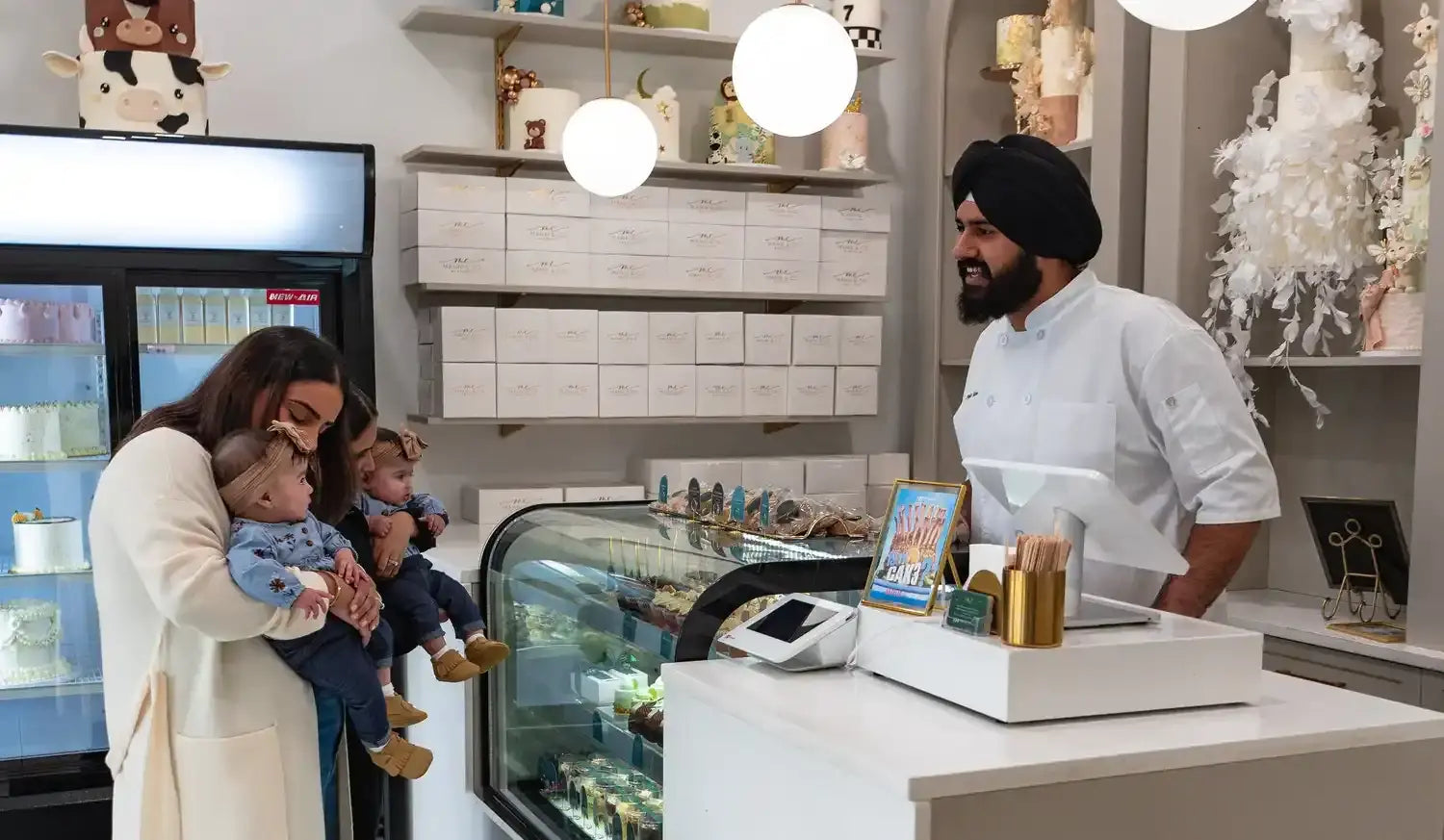 Woman holding a baby and a toddler in a bakery, with a baker behind the counter.